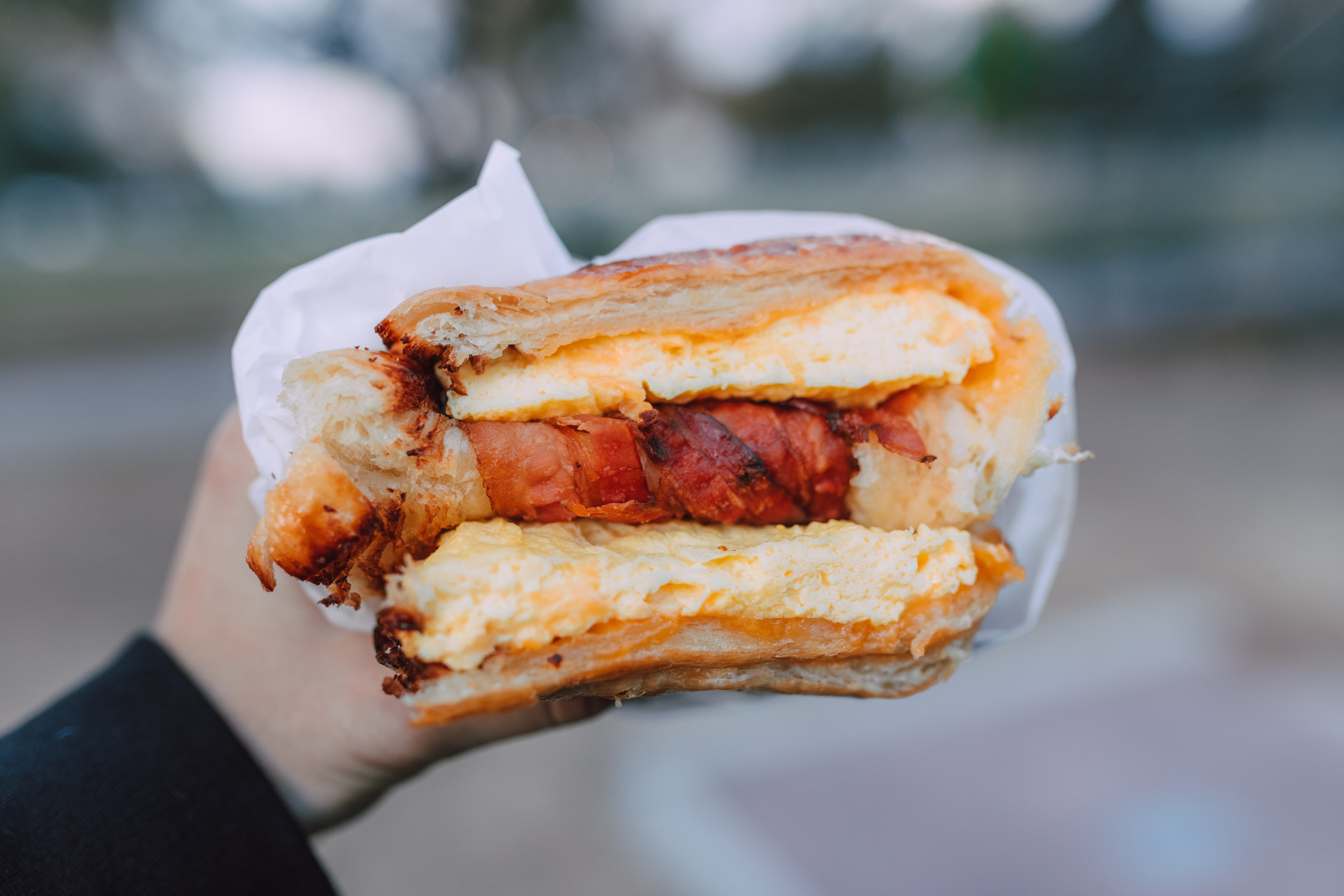 Hand holding a bacon, egg, and cheese sandwich while outdoors.