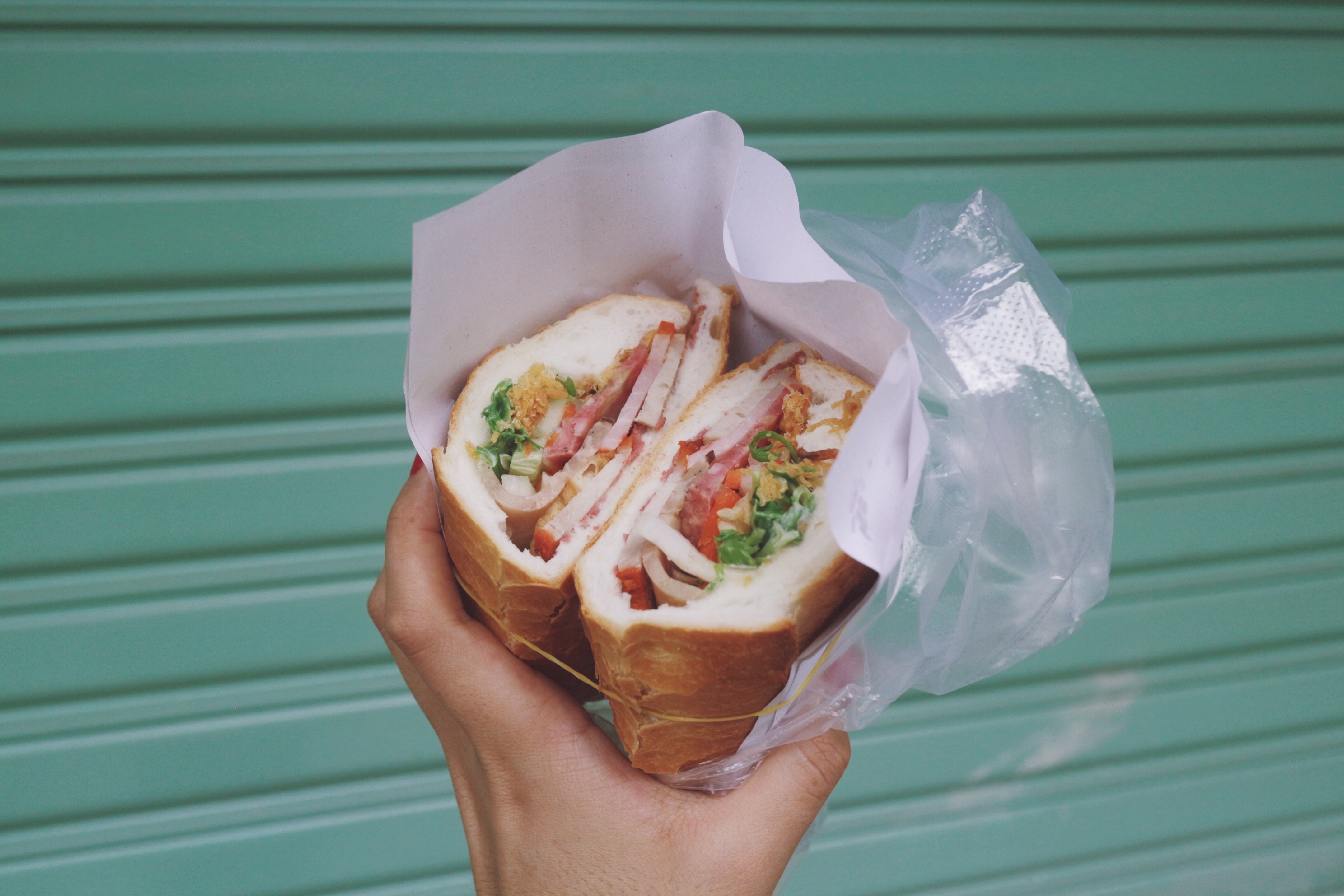 Hand holding a Cuban sandwich with ham, tomatoes, and lettuce in front of a teal gate.