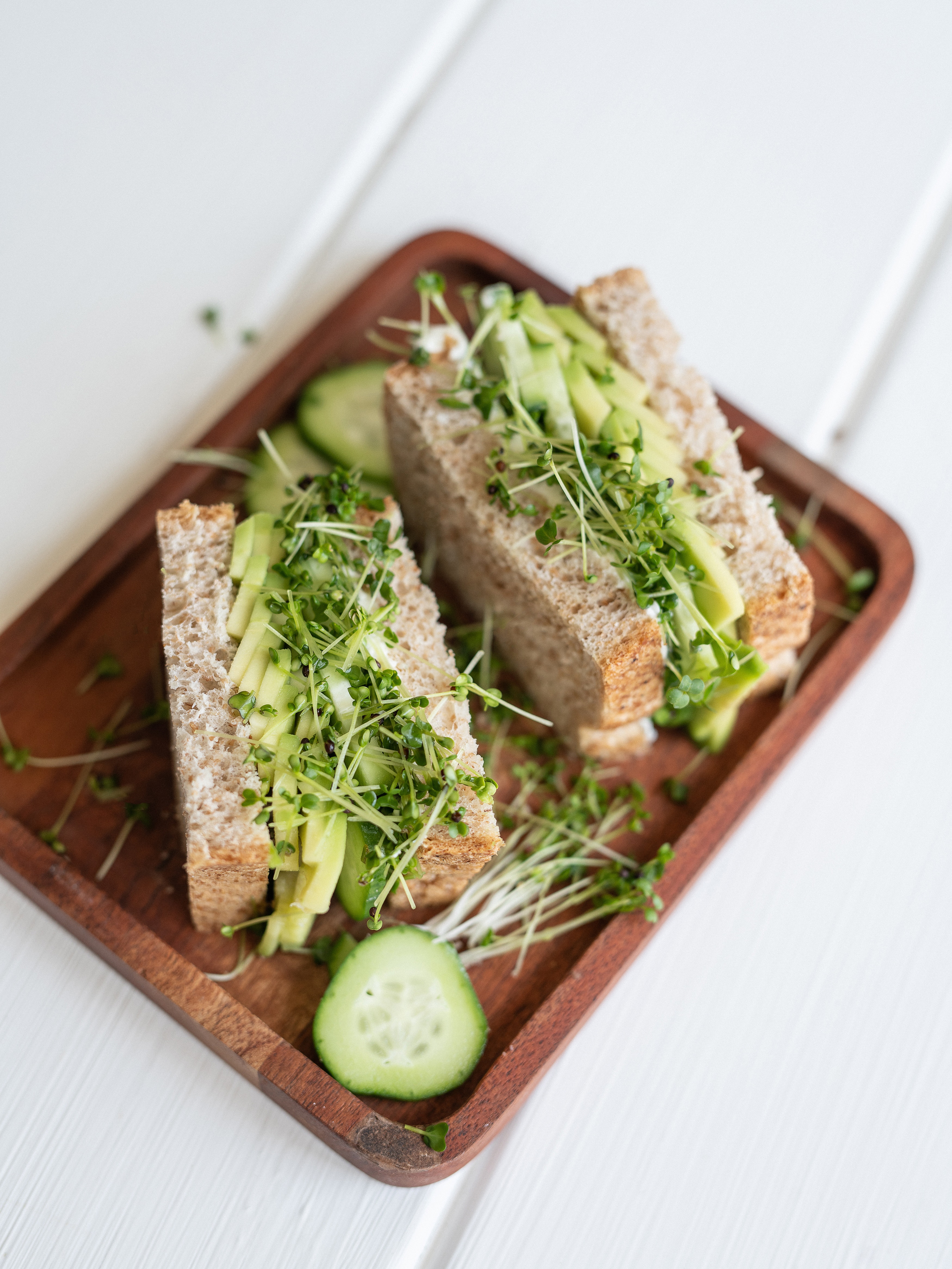 Sandwich with cucumber, avocado, and microgreens on a wooden tray.